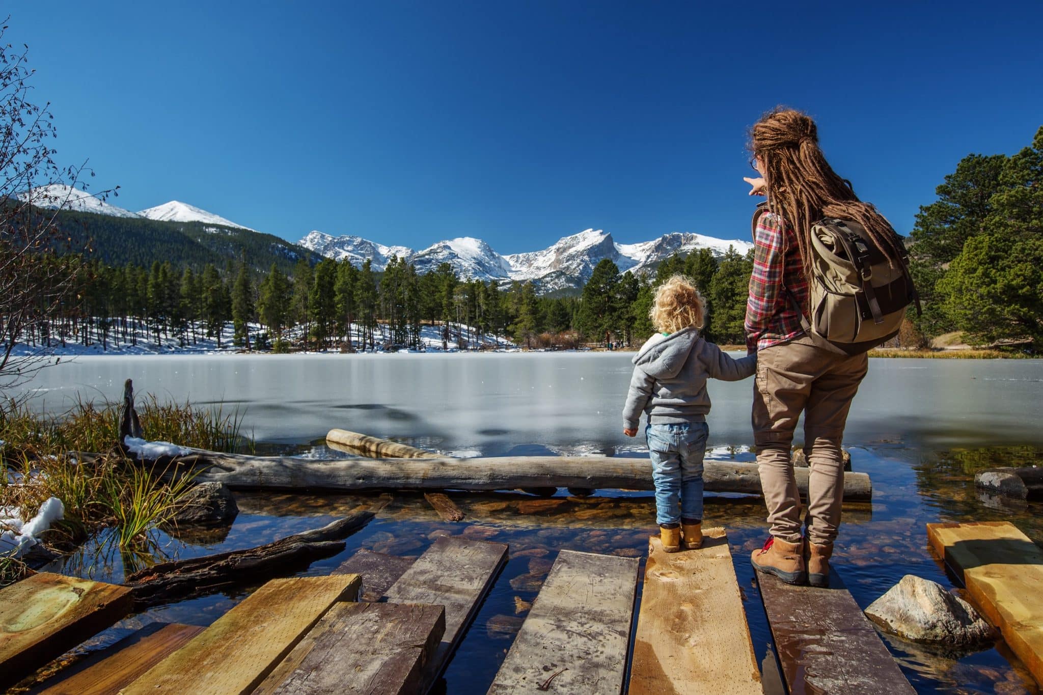 Adult and child holding hands on a wooden dock, facing a tranquil mountain lake, depicting the serenity sought by families awaiting the Canadian spouse visa processing time 2024.
