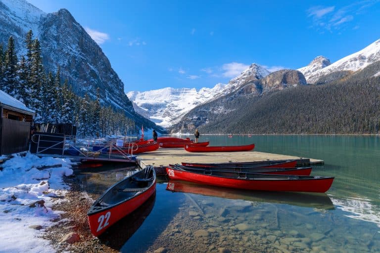 Visit Canada to see red canoes lined up on the shore of a snow-dusted lake with mountains in the background.