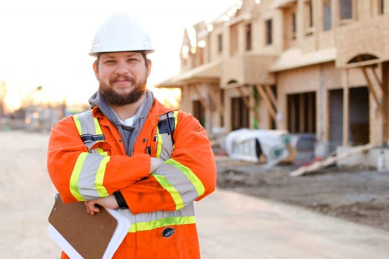 Foreman in orange workwear holding notebook on construction site, house in background. Concept of real estate in Canada.