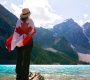 A person, who recently completed their UK to Canada immigration journey, draped in a Canadian flag stands on a lakeshore looking at mountainous scenery.