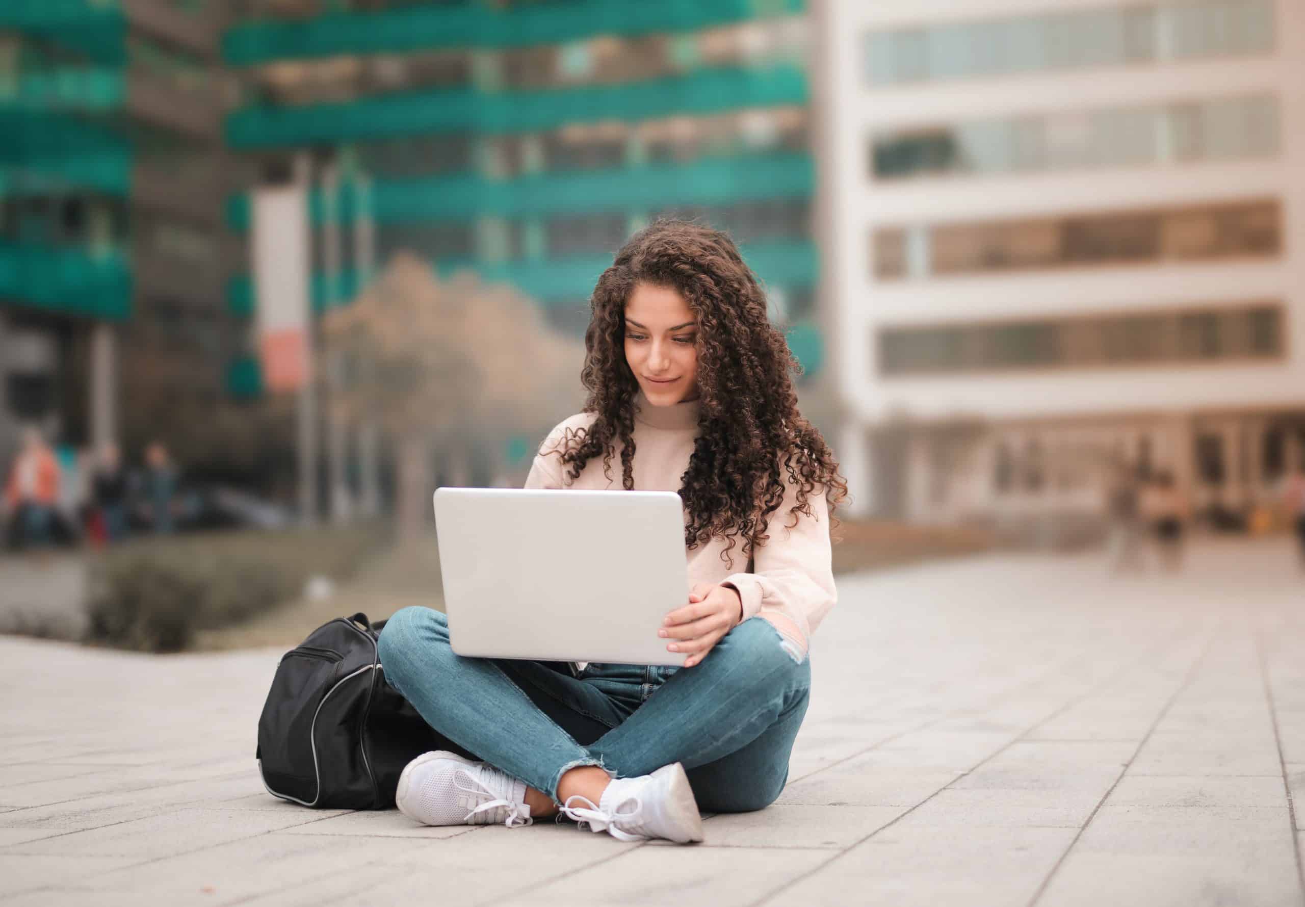 A young woman sitting cross-legged on the ground using a laptop with buildings in the background, working on her Canada study permit application.