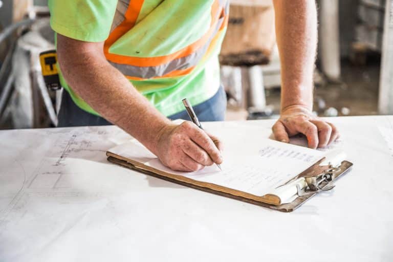 A Canada Federal Skilled Worker in construction reviews plans and takes notes on a clipboard.