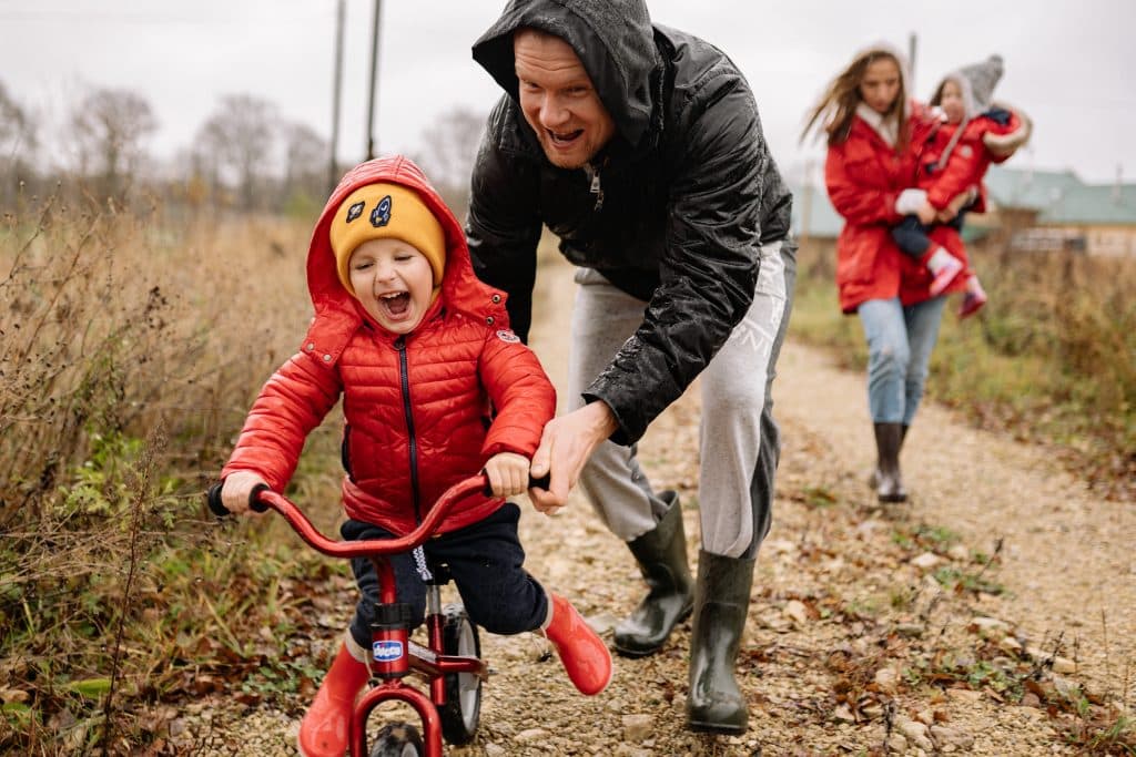 A child learning to ride a bike with the help of an adult while another person watches in the background during a Canadian family sponsorship gathering.