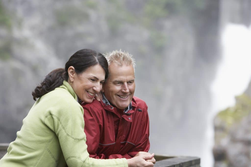 A man and woman stand close together, leaning on a railing, smiling while looking off-camera. A waterfall and rocky scenery are blurred in the background, capturing a perfect moment that symbolizes their hopes for a Canada spouse visa 2024.